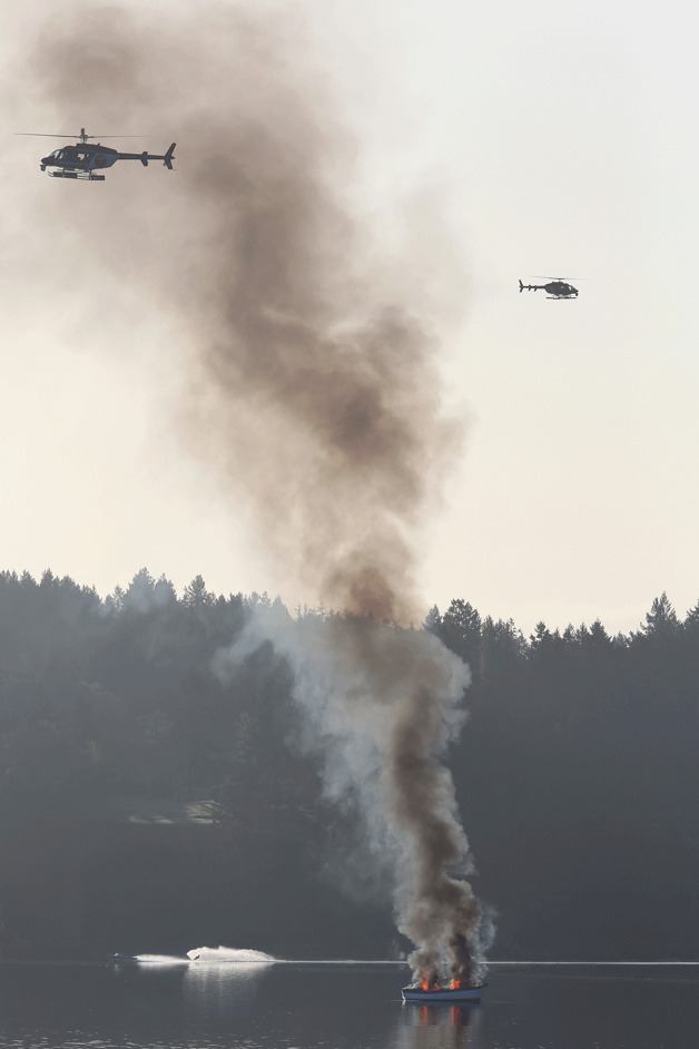 A boat in Agate Pass goes up in flames as news helicopters hover overhead and a boat from Bainbridge Island motors past. In the boat is driver Jim Llewellyn pulling skier Scott Norrie.